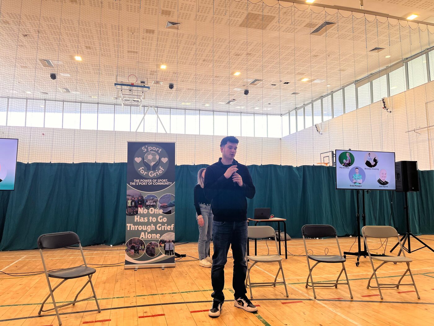 A speaker addressing students in a sports hall beside a S'port for Grief banner